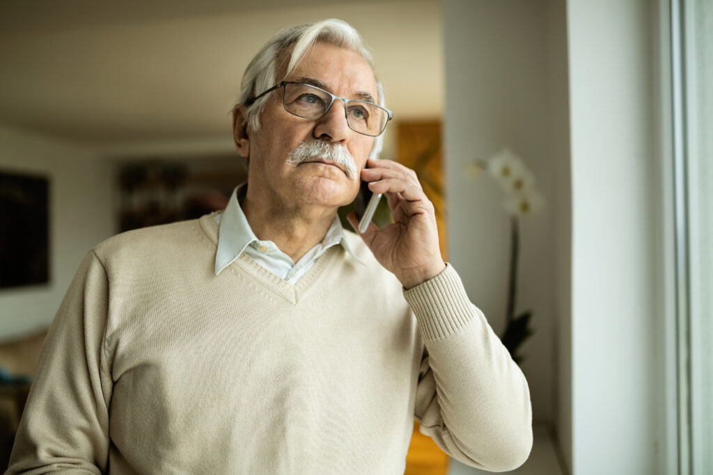 Senior man with glasses and a beige sweater speaking on a mobile phone in a well-lit room with a window.