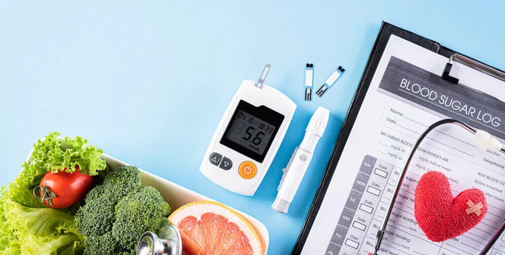 Blood sugar monitor, fresh vegetables, & a blood sugar log on a table.