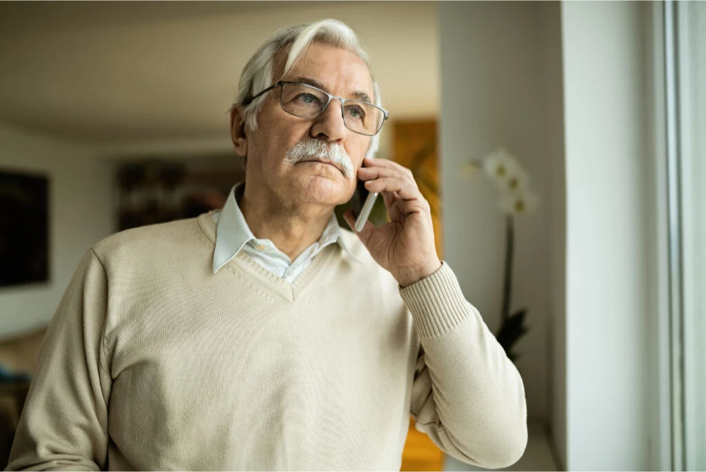 Elderly man with glasses talking on a phone near a window.