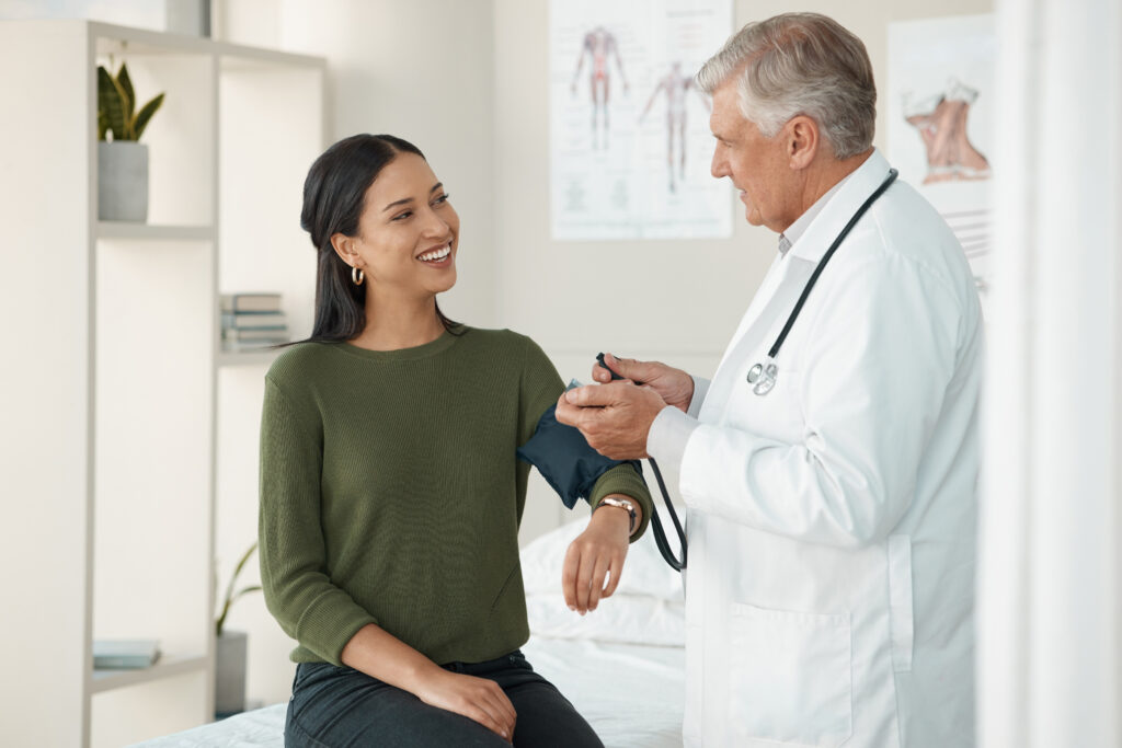 Doctor checking blood pressure of a smiling female patient in a well-lit clinic room.