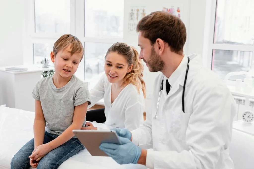 Doctor with a stethoscope around his neck showing a tablet to a young boy and his mother in a bright medical office.