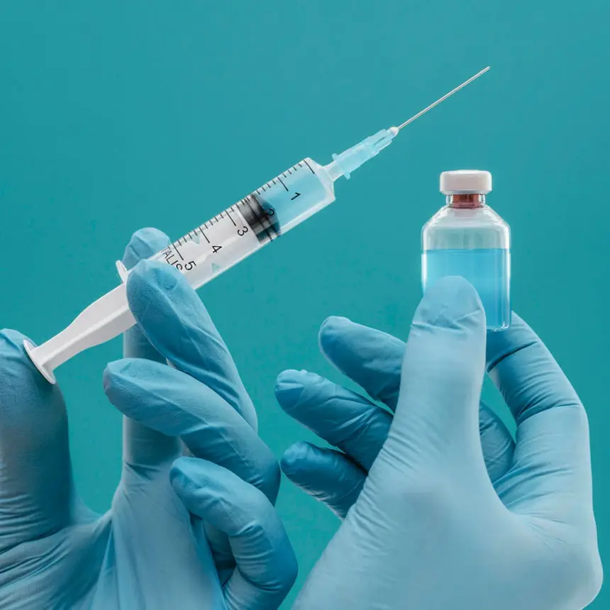 Hand in blue gloves holding a syringe and vaccine vial against a turquoise background.