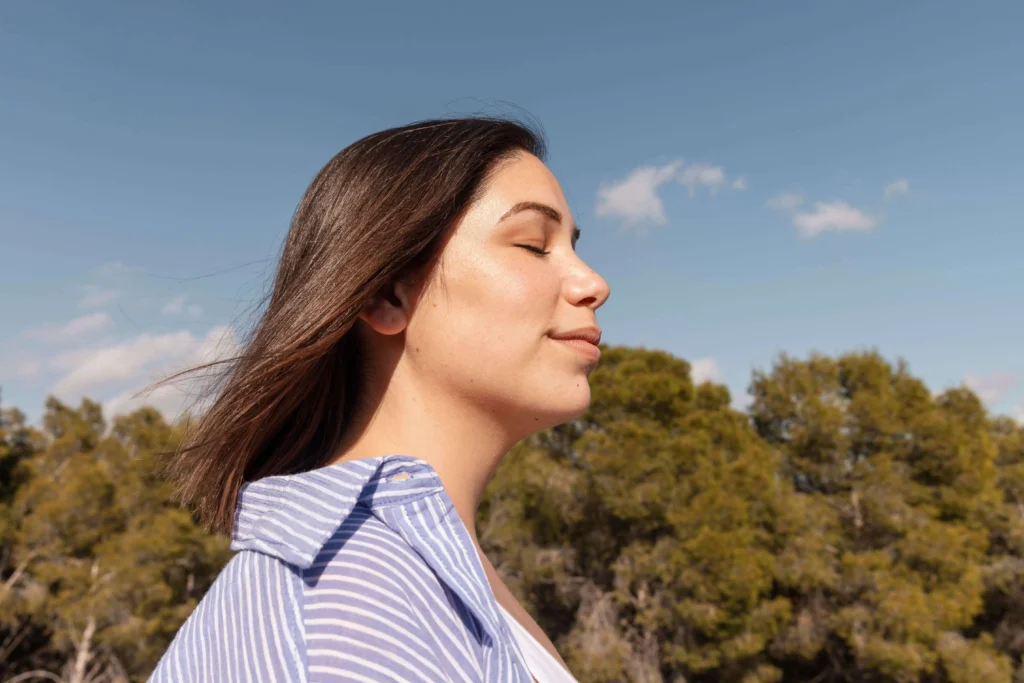 A woman with her eyes closed, smiling and breathing in fresh air outdoors with trees in the background.