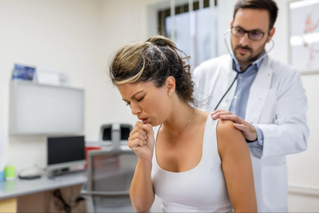 A woman is coughing while a doctor listens to her lungs with a stethoscope in a medical office.