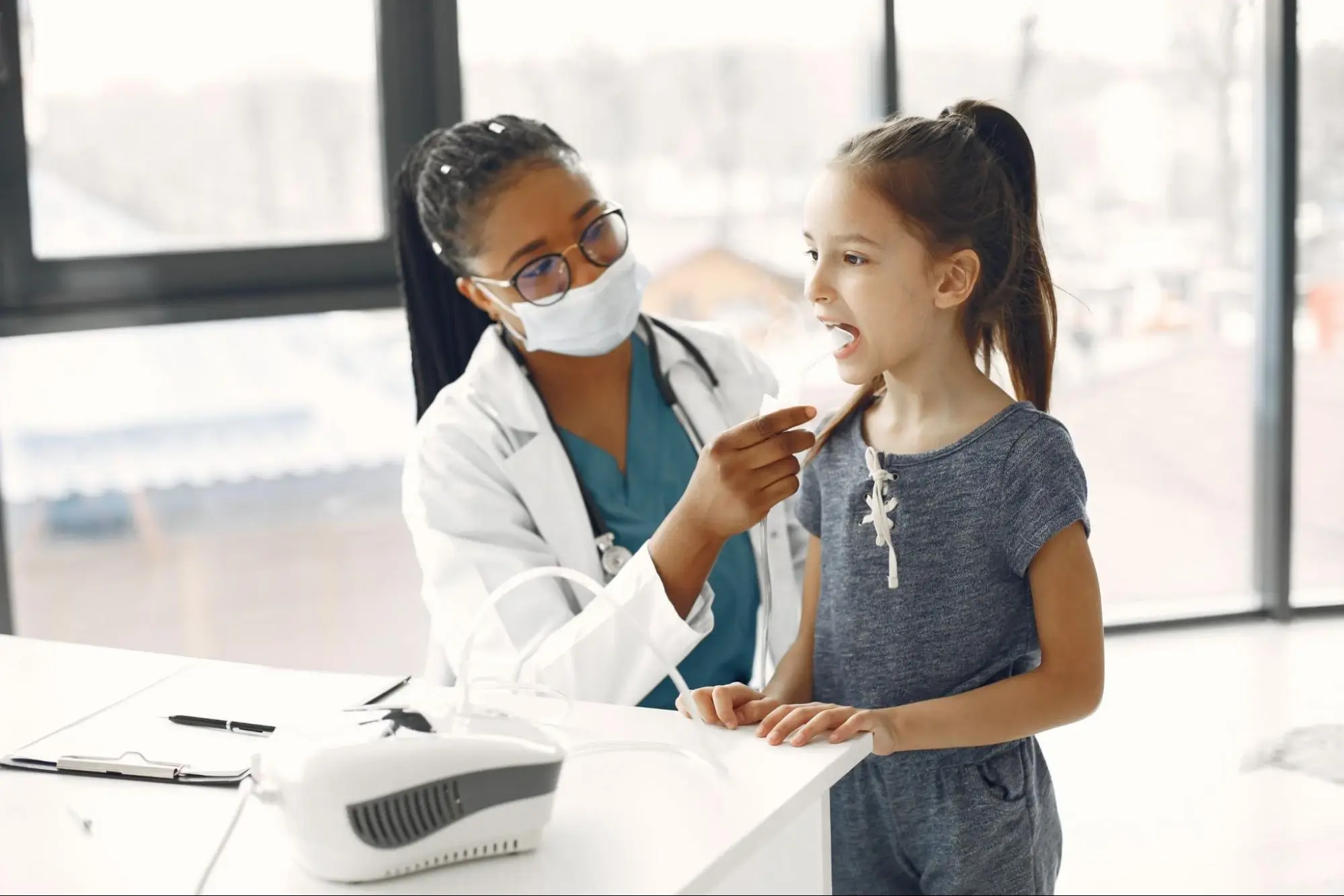 A doctor wearing a mask and stethoscope uses a tongue depressor to examine a young girl's throat in a brightly lit medical office.