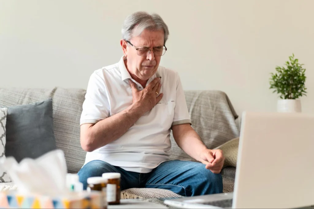 An older man sitting on a couch, holding his chest in discomfort while looking at a laptop.