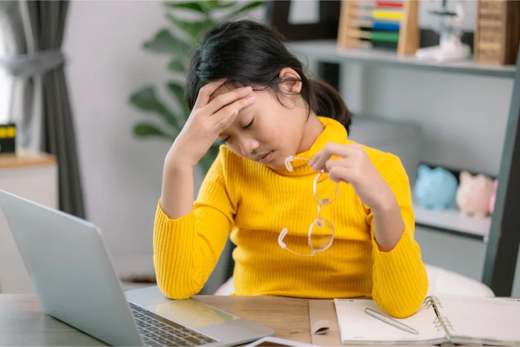 A young girl sitting at a desk, rubbing her forehead and holding her glasses, looking tired.