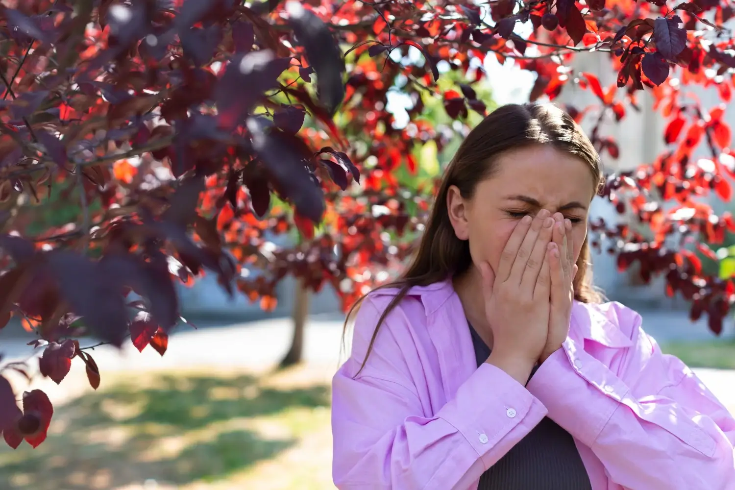 A woman standing near red leaves outdoors, covering her nose while sneezing.