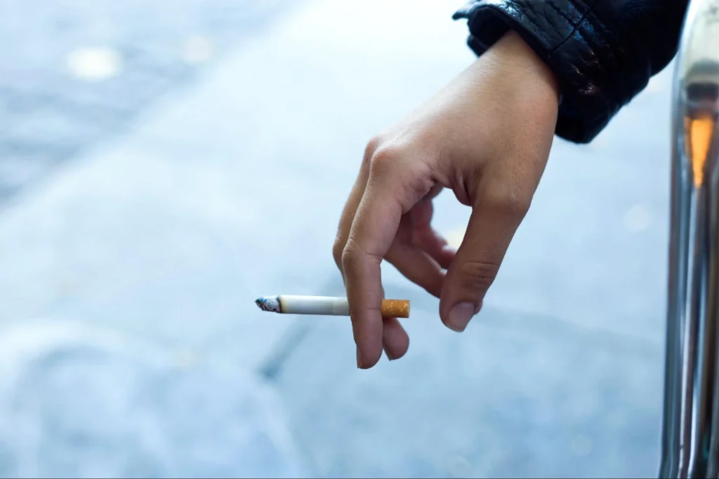 A close-up of a hand holding a cigarette, with a street background.