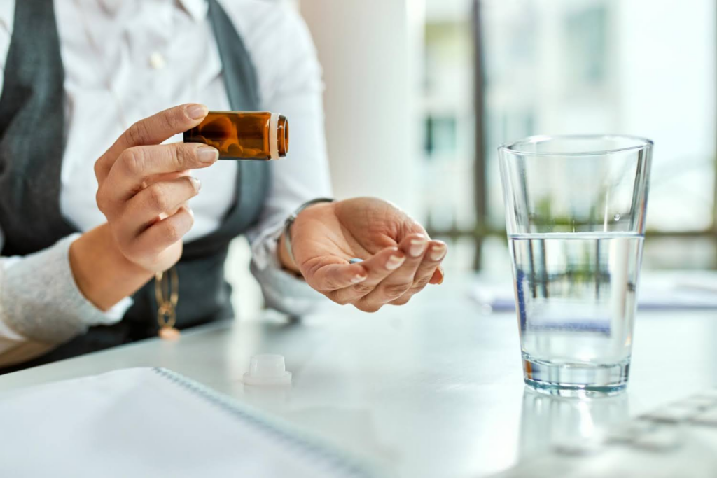 A woman holding a pill in one hand and a bottle in the other, with a glass of water on the desk.