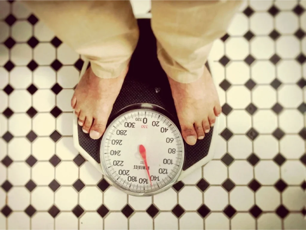 A person standing on a weight scale with a view of the dial, showing weight in pounds.