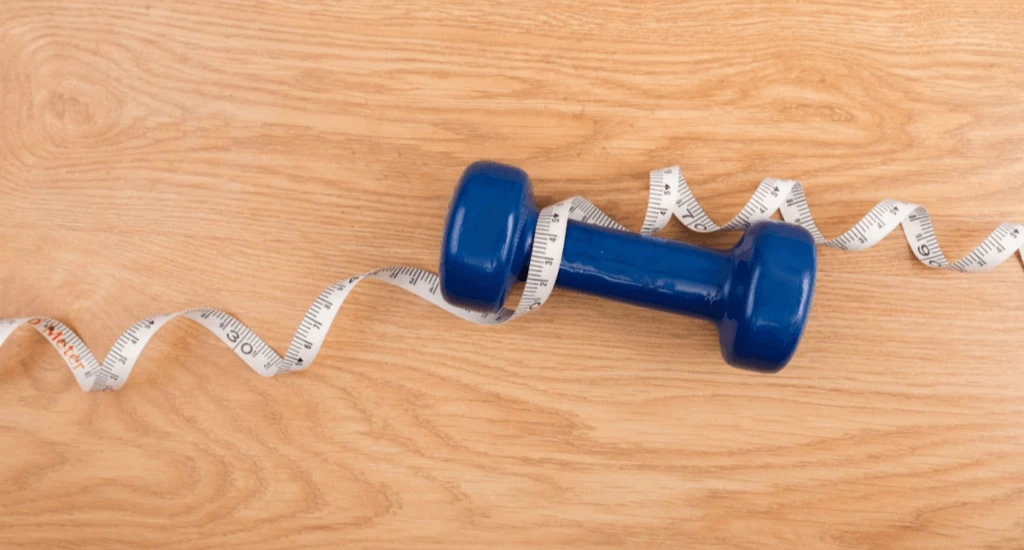 A blue dumbbell placed on a wooden surface with a measuring tape wrapped around it.