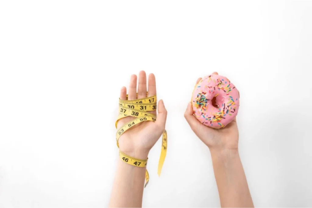 Two hands, one holding a yellow measuring tape and the other holding a pink frosted donut.