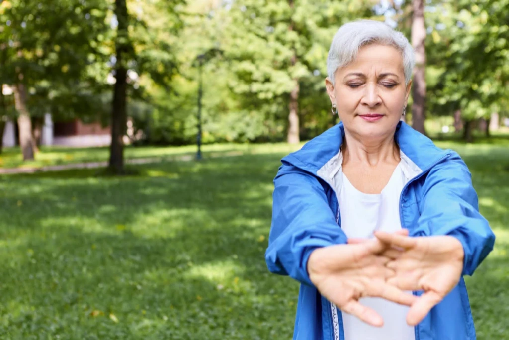 An elderly woman performing hand stretches in a park, wearing a blue jacket.