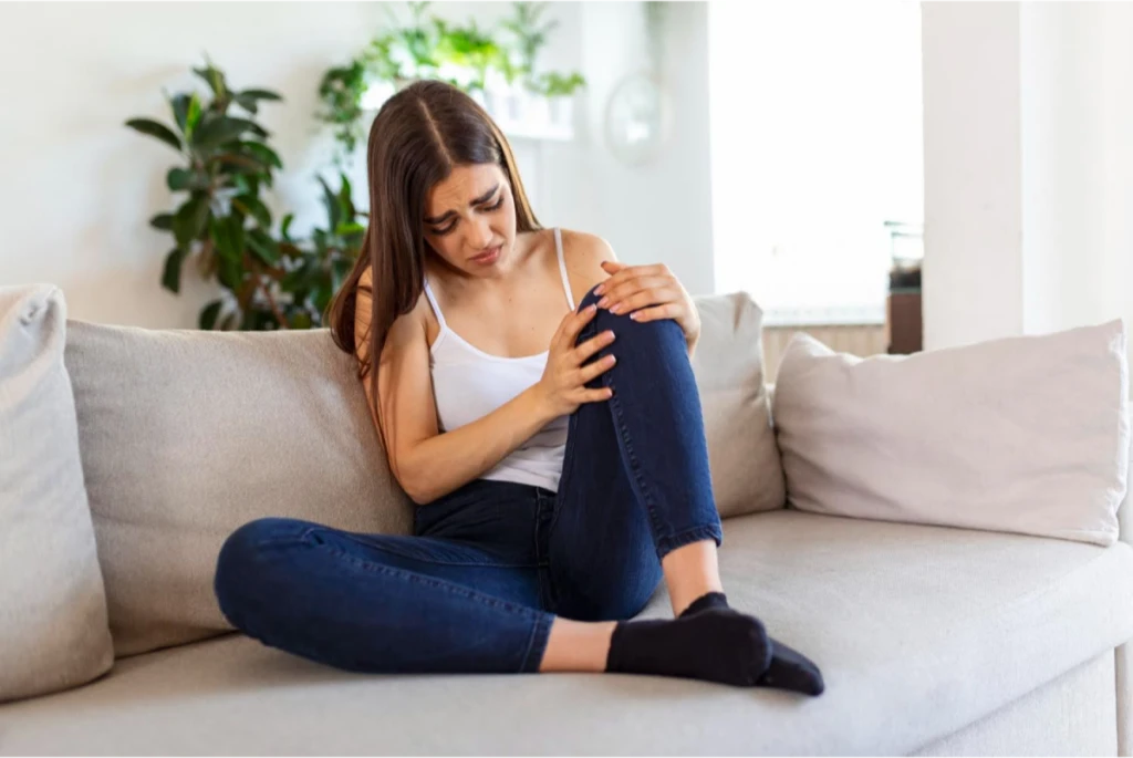 A young woman sitting on a couch, holding her knee in pain.